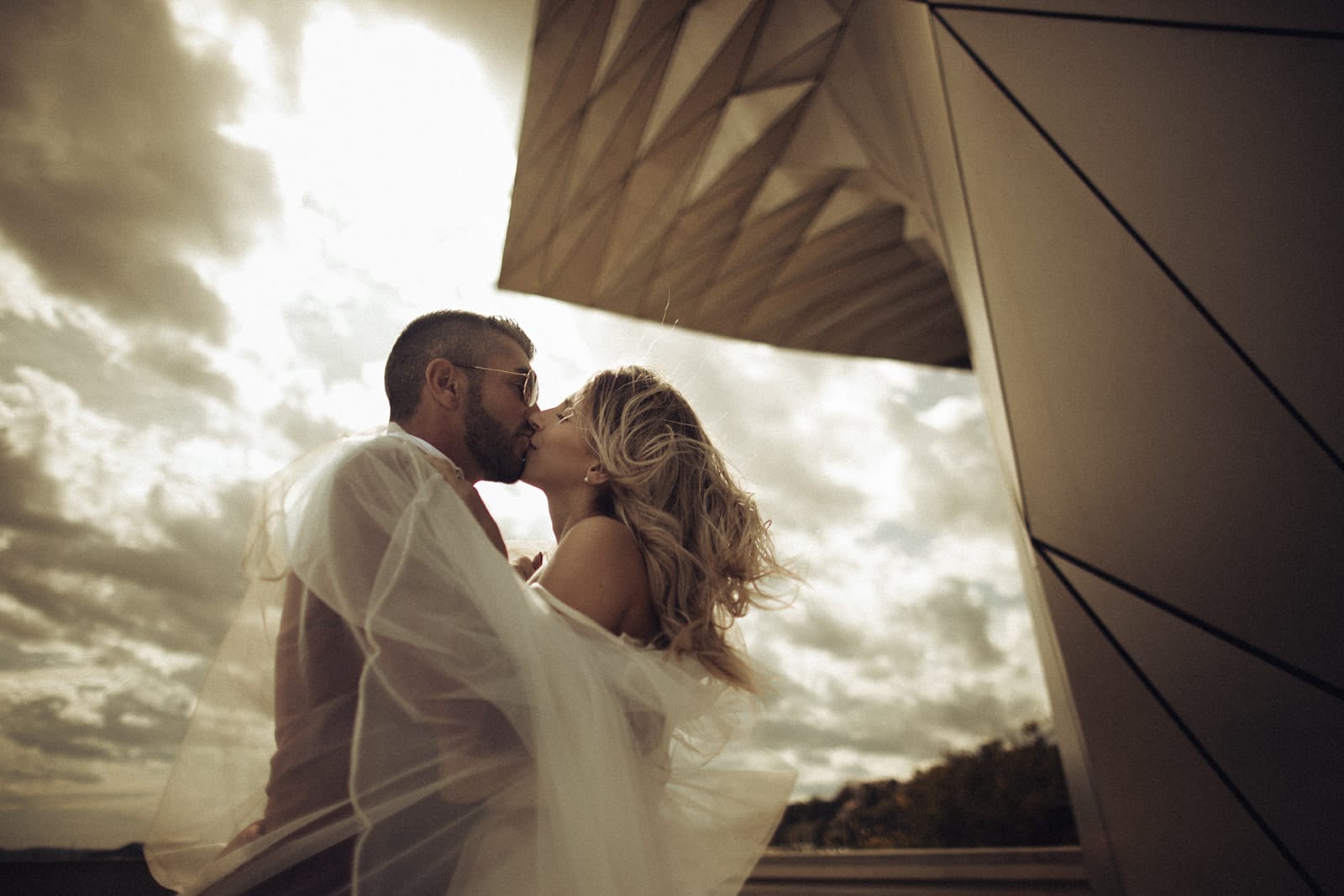 Gros plan d’un baiser entre deux mariés sur un rooftop, enveloppés dans le voile, avec une architecture contemporaine en arrière-plan. Par Castille Alma photographe de mariage à Lyon et Bangkok