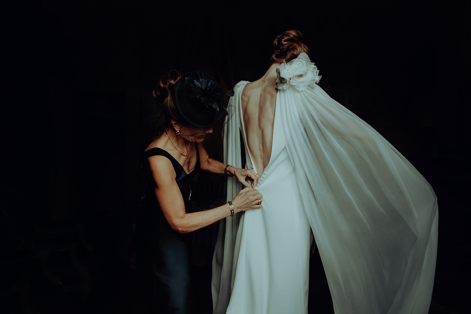 photographe mariage Lyon @castillealma La mère de la mariée aide la mariée à fermer les derniers boutons d’une robe dos nu aux détails vaporeux, dans une lumière feutrée et intime – par Castille Alma, photographe de mariage en France et Bangkok
