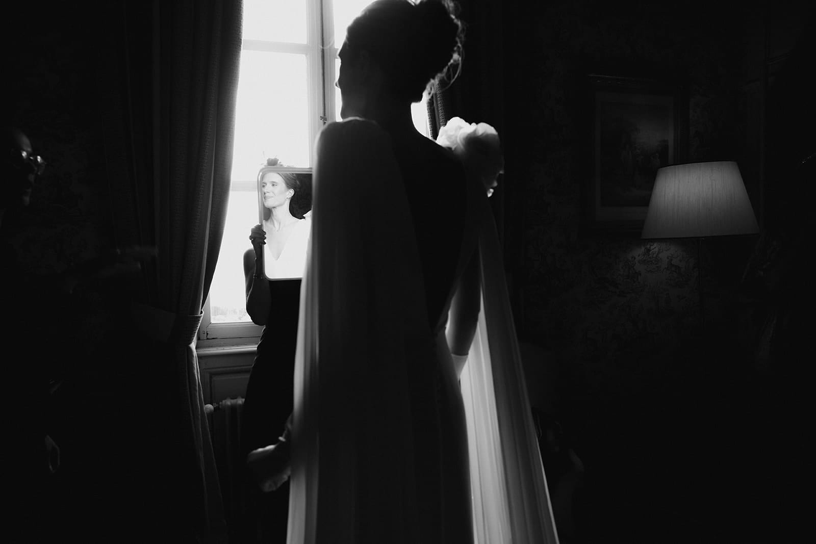 photographe mariage Lyon @castillealma La mariée découvre son reflet dans un miroir tenu à bout de bras devant une fenêtre, lumière douce sur son visage, ombres autour d’elle – par Castille Alma, photographe de mariage en France