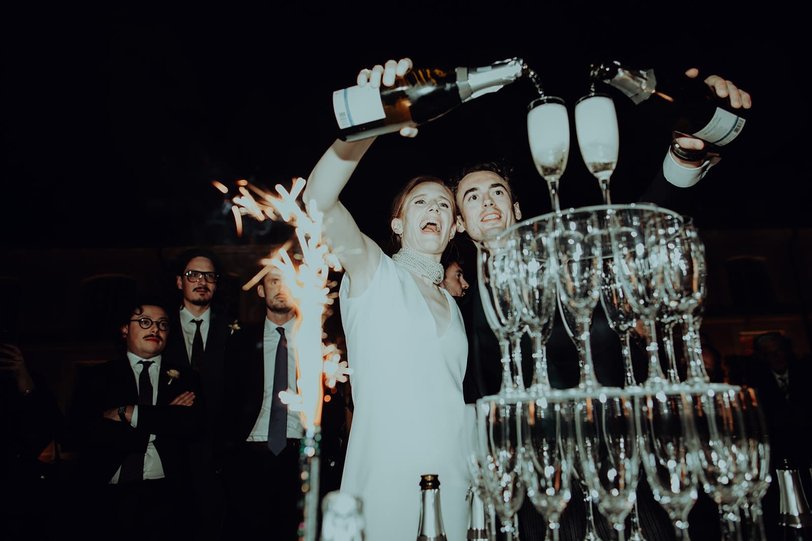 Les mariés versent le champagne en cascade, visages illuminés par les feux de bengale, éclats de voix et de bulles autour d’eux – par Castille Alma, photographe de mariage à Phuket et partout en Asie