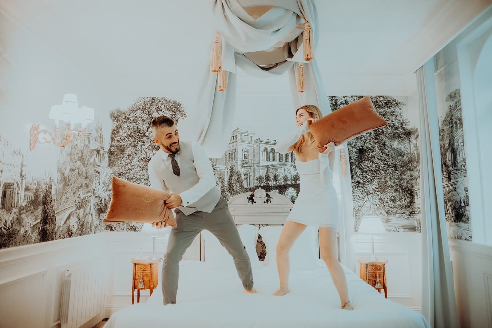 galerie photographe mariage Lyon @castillealma Un couple de jeunes mariés joue à la bataille d’oreillers sur un lit à baldaquin, rires complices et déco royale autour – par Castille Alma, photographe de mariage à Zurich, Genève et partout en Europe