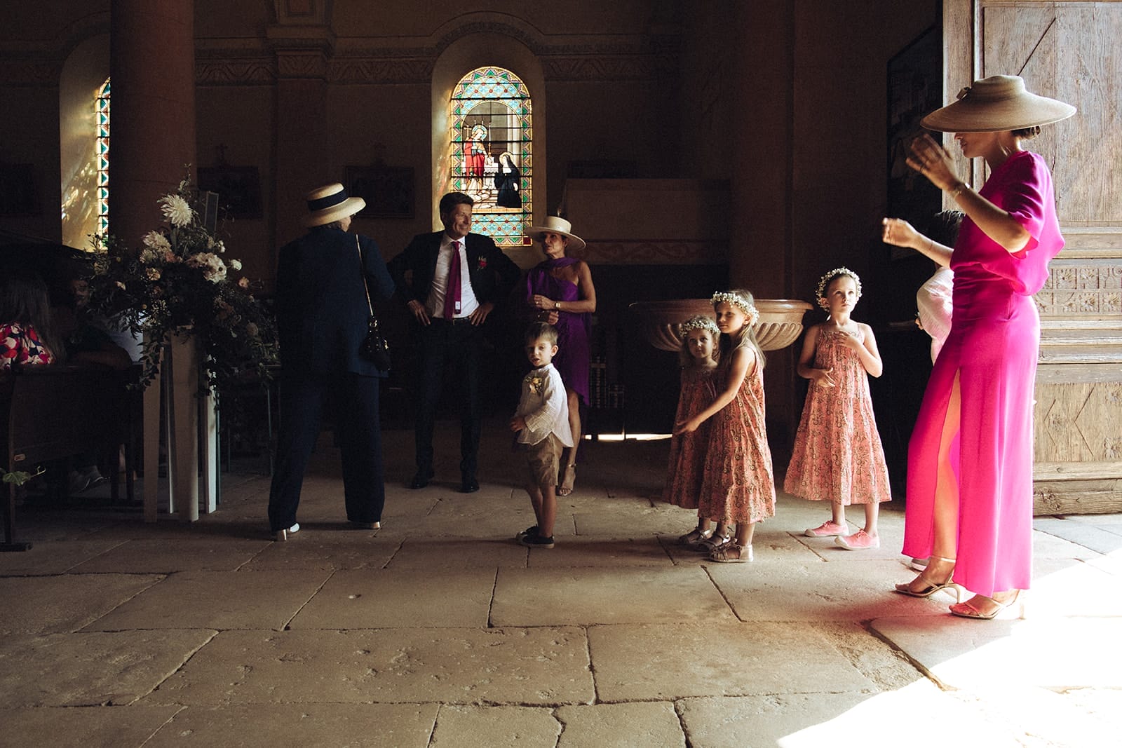 photographe mariage Lyon @castillealma Scène d’attente avant une cérémonie religieuse : enfants d’honneur en ligne, adultes chapeautés, éclats de voix et lumière rasante dans une église – par Castille Alma, photographe de mariage en France