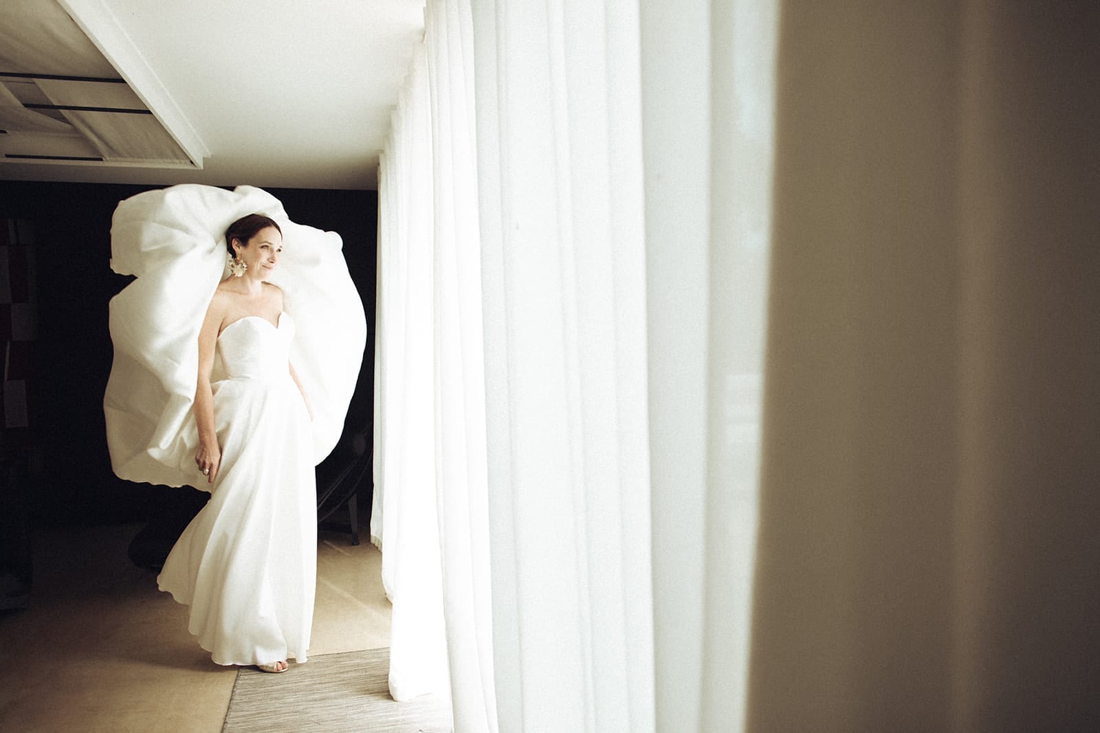 photographe mariage Lyon @castillealma Une mariée lumineuse dans sa robe sculpturale, debout face à la lumière des rideaux blancs, quelques secondes avant de dire oui – par Castille Alma, photographe de mariage en FranceLyon Bangkok
