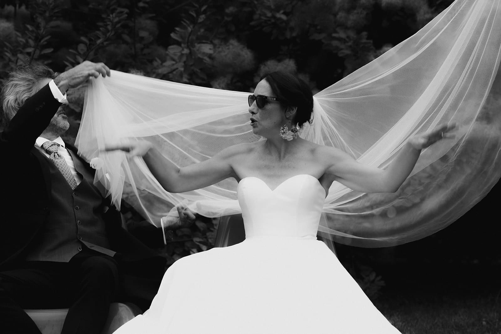 galerie photographe mariage Lyon @castillealma Castille Alma photographe de mariage à Lyon Bangkok la mariée rock reajuste son voile Une mariée en robe bustier blanche joue avec son voile tenue par un homme plus âgé, dans une scène vivante et complice en extérieur – par Castille Alma, photographe de mariage en Europe