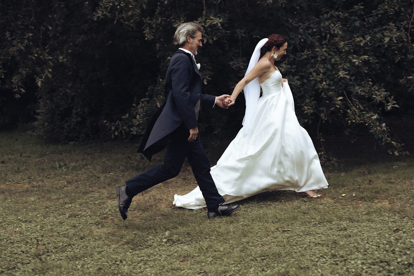 Un couple de mariés court main dans la main sur l’herbe, robe au vent et sourires complices, instant pris sur le vif – par Castille Alma, photographe de mariage en Europe