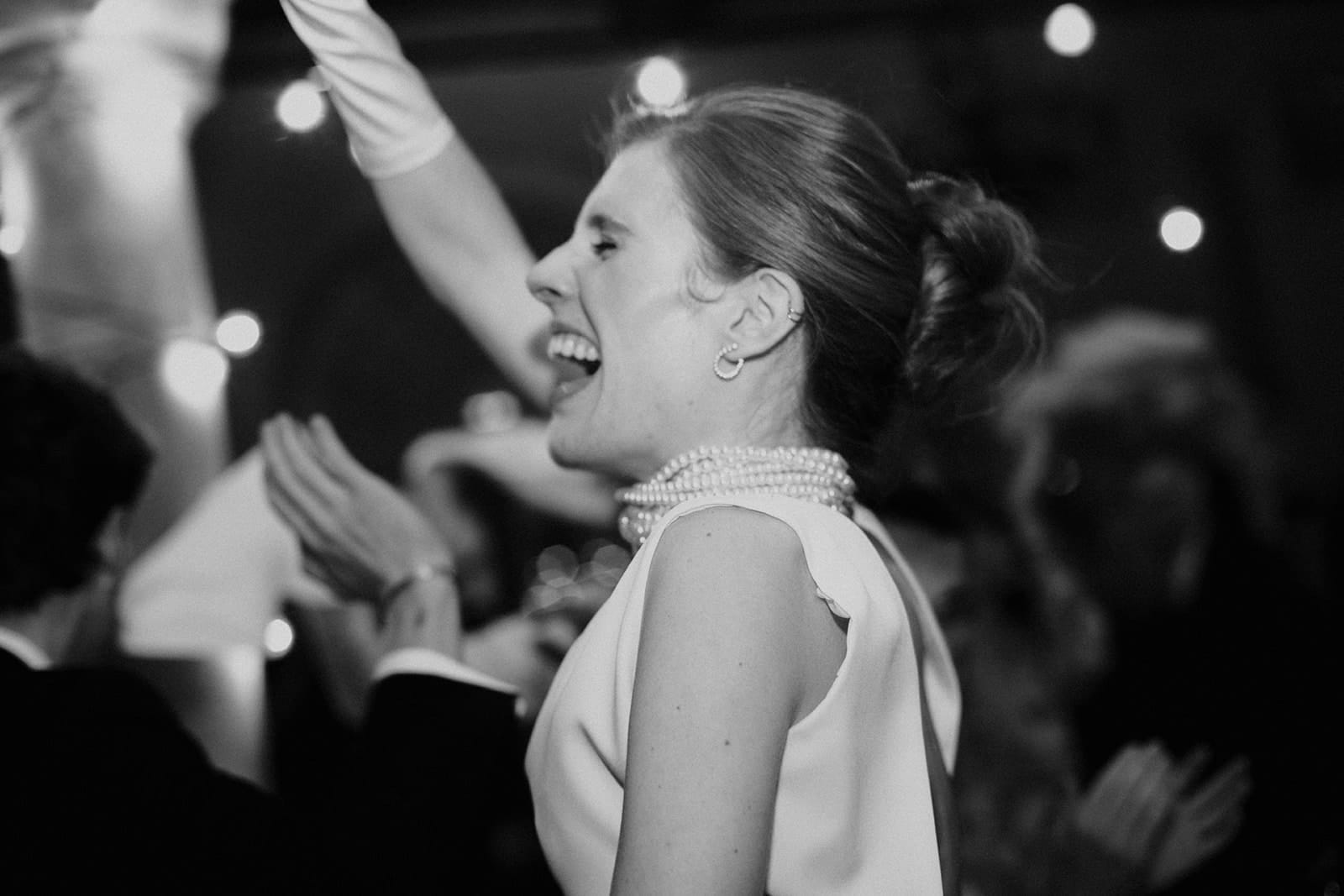 galerie photographe mariage Lyon @castillealma La mariée éclate de rire en pleine soirée, perles au cou, bras levés, lumière douce et ambiance électrique autour – par Castille Alma, photographe de mariage à Chiang Mai et partout en Asie