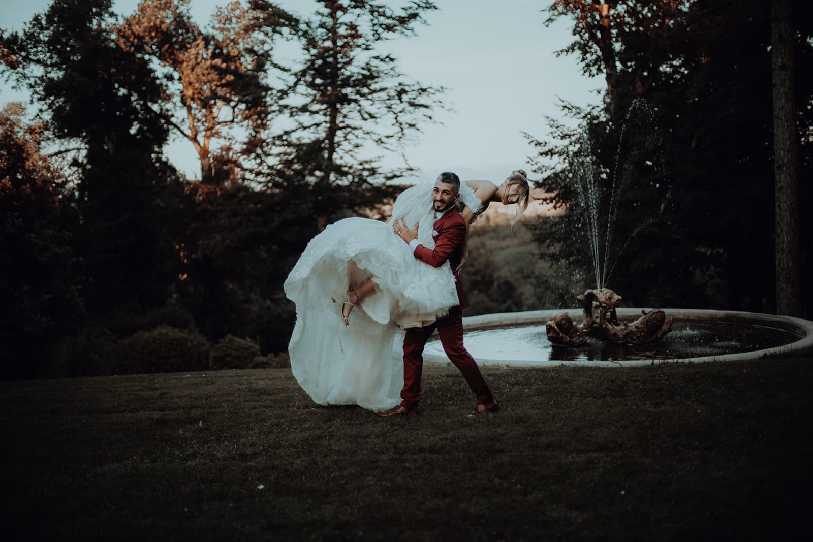 galerie photographe mariage Lyon @castillealma Castille Alma photographe de mariage à Lyon Bangkok Le marié porte la mariée sur son epaule comme un enlevement. Un marié en costume bordeaux porte sa femme en robe de princesse devant une fontaine, tous deux rayonnants dans un décor de nature en fin de journée – par Castille Alma, photographe de mariage en France, en Europe, à Bangkok ou ailleurs.