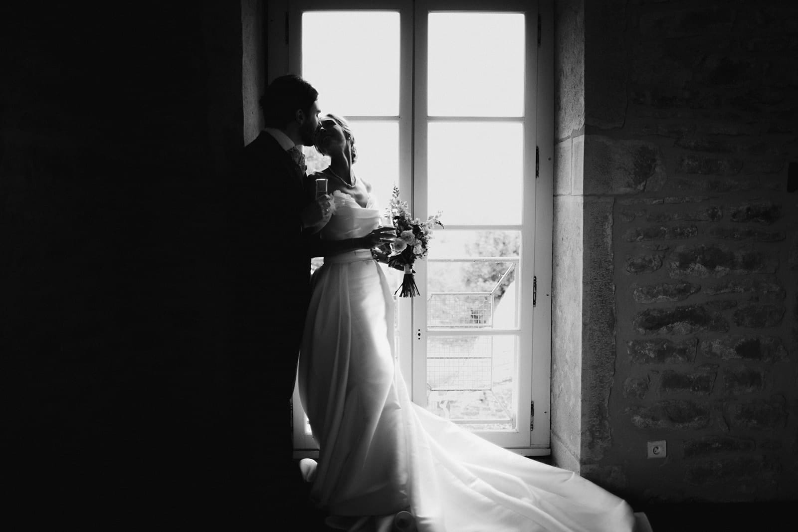 Les mariés s’embrassent à contre-jour devant une fenêtre ancienne du Château de Pierreclos – photo signée Castille ALMA