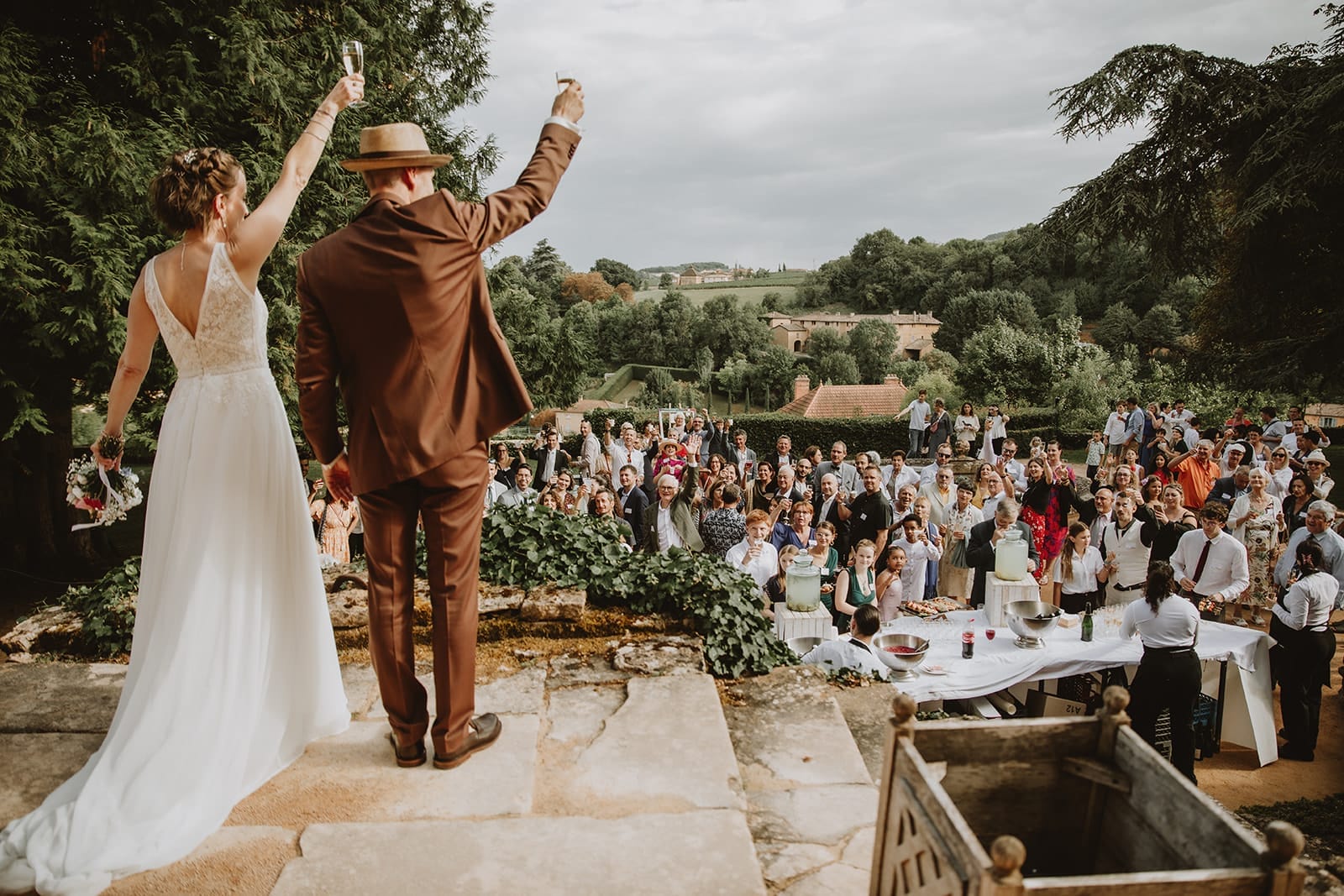 Mariés trinquant au cocktail de leur mariage au Manoir de la Garde à Ville-sur-Jarnioux, avec vue sur les invités et les collines du Beaujolais