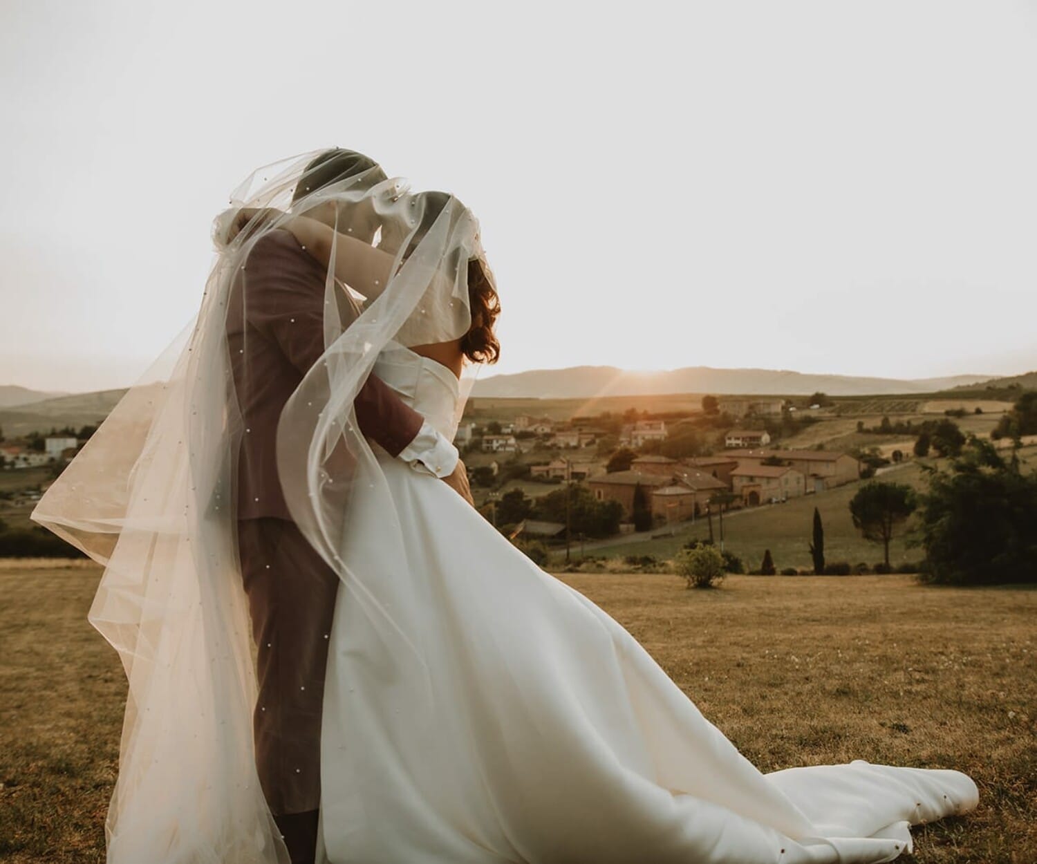 Mariés enlacés sous un voile, lumière dorée et village en arrière-plan, lors d’un mariage musical à la Bastide des Hirondelles.