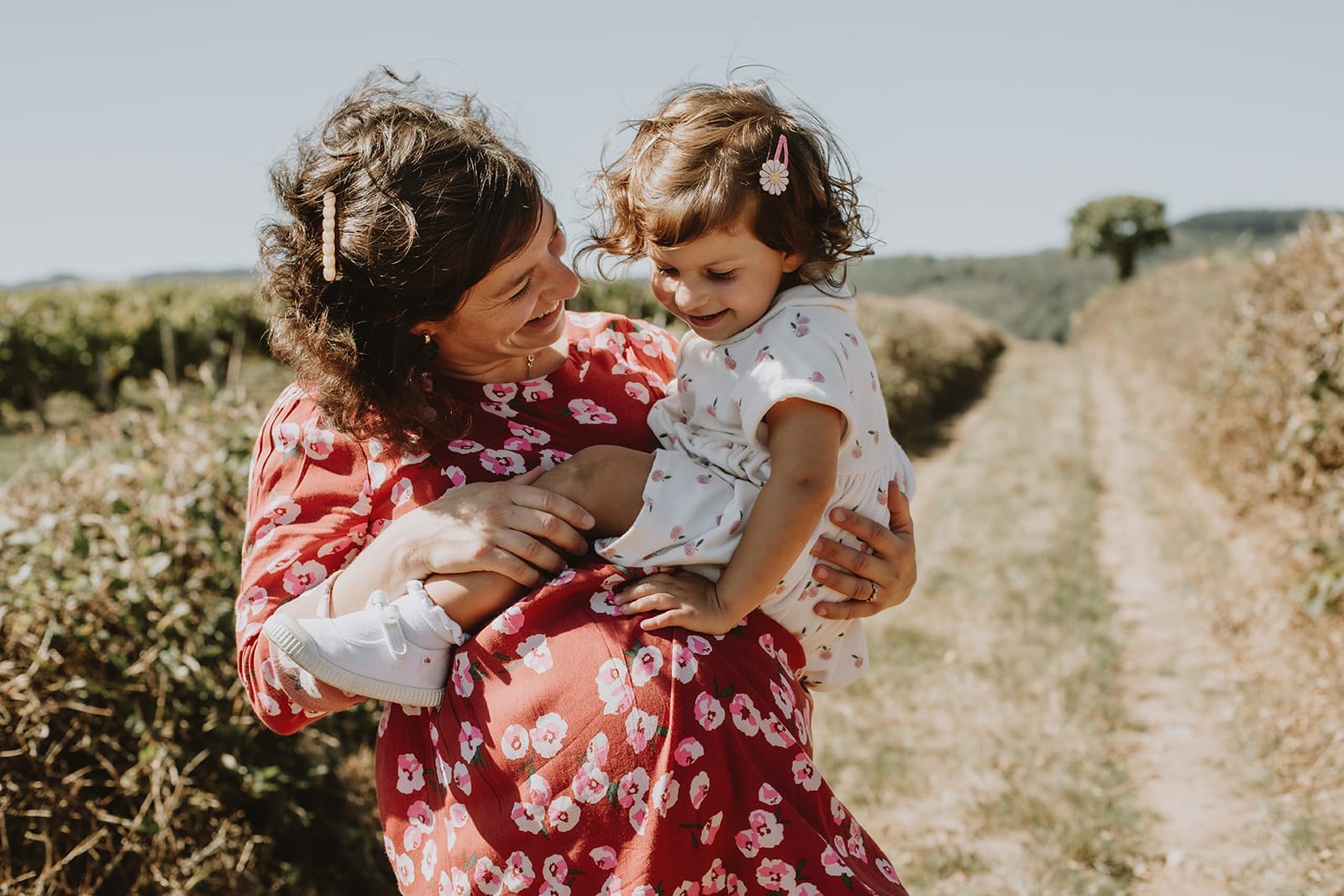 Une maman souriante tient sa petite fille dans un champ ensoleillé.