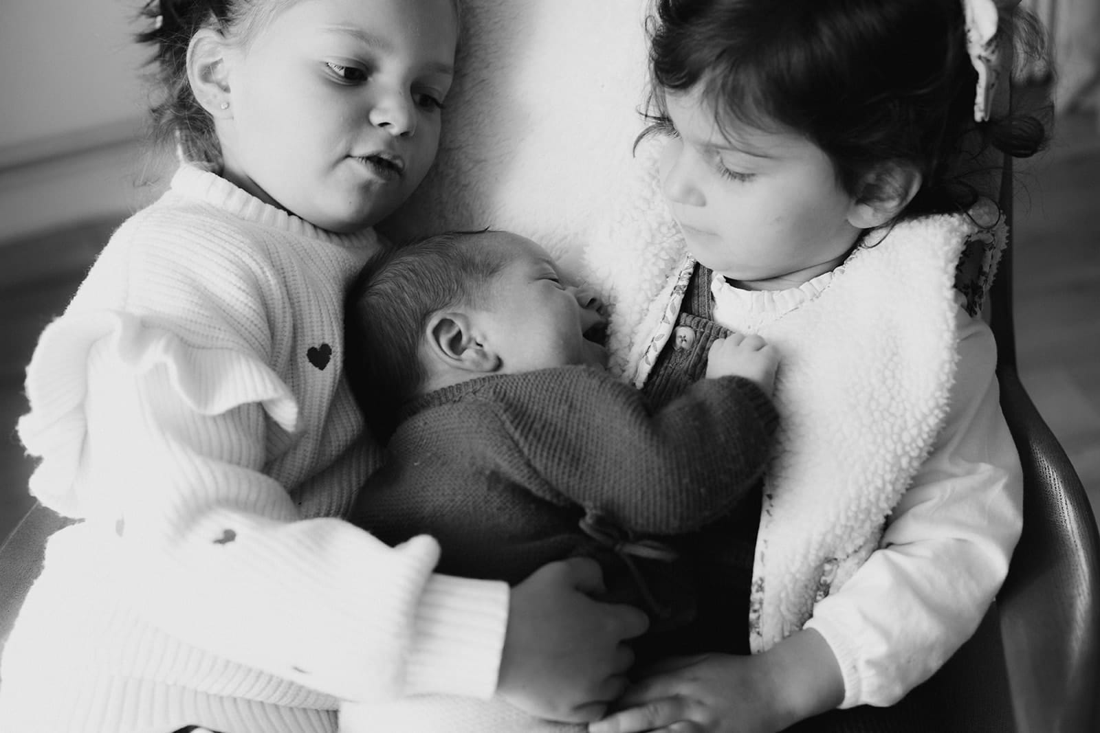 Deux petites filles entourent un bébé souriant, créant un moment tendre.